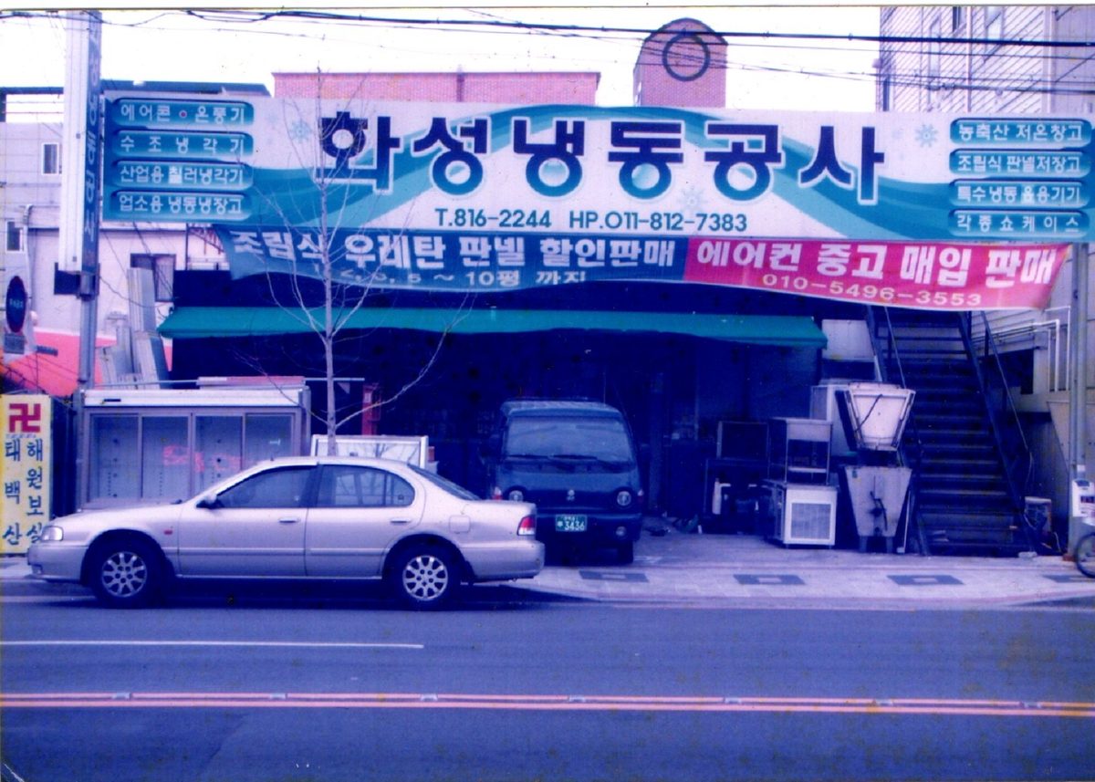 The original Hwasung Refrigeration workshop in Korea. Its signage reads
                      Hwasung Refrigeration Company in Korean, with cold-storage units and
                      insulated panels stacked outside.